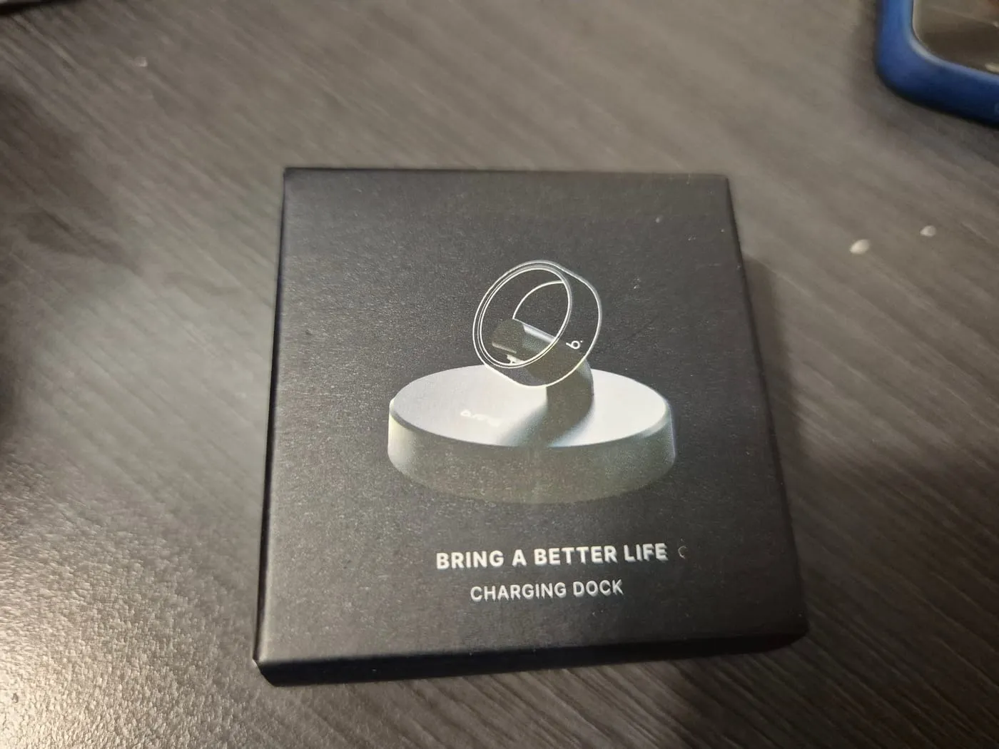 b.ring 상자. 검정 무광 표면에 충전 독 이미지와 BRING A BETTER LIFE, CHARGING DOCK 문구가 있다