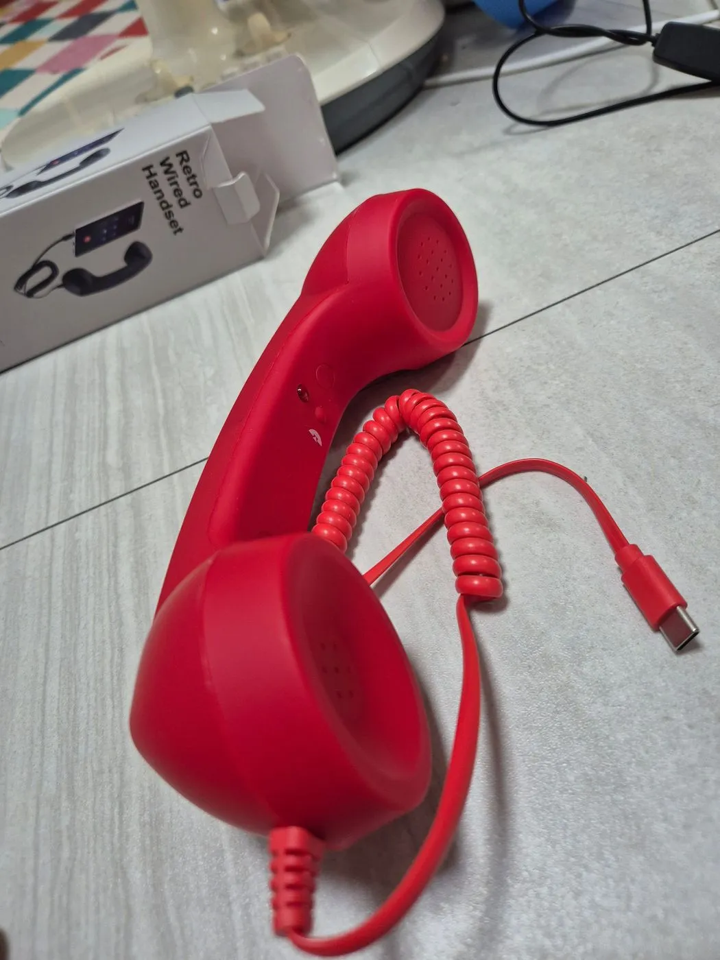 빨간 레트로 수화기와 코일 케이블, 끝의 USB-C 단자가 바닥에 놓여 있다. 뒤로 상자가 보인다